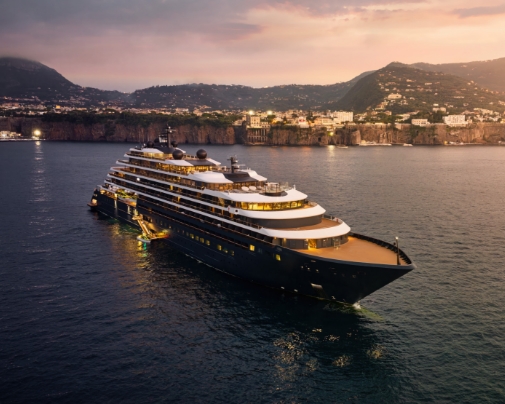 A luxury yacht anchored off a coastline at golden hour with mountains in the distance.